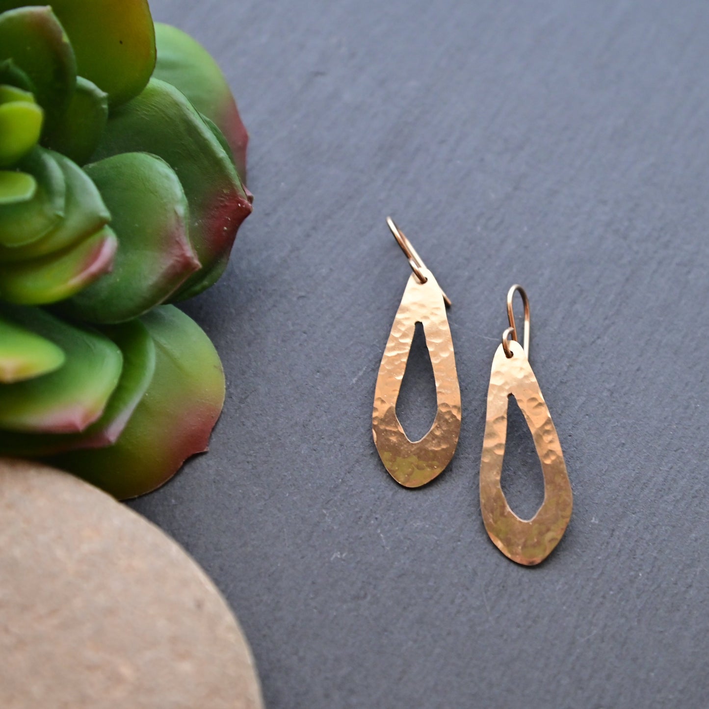 Gold teardrop earrings on a dark surface with a succulent plant.