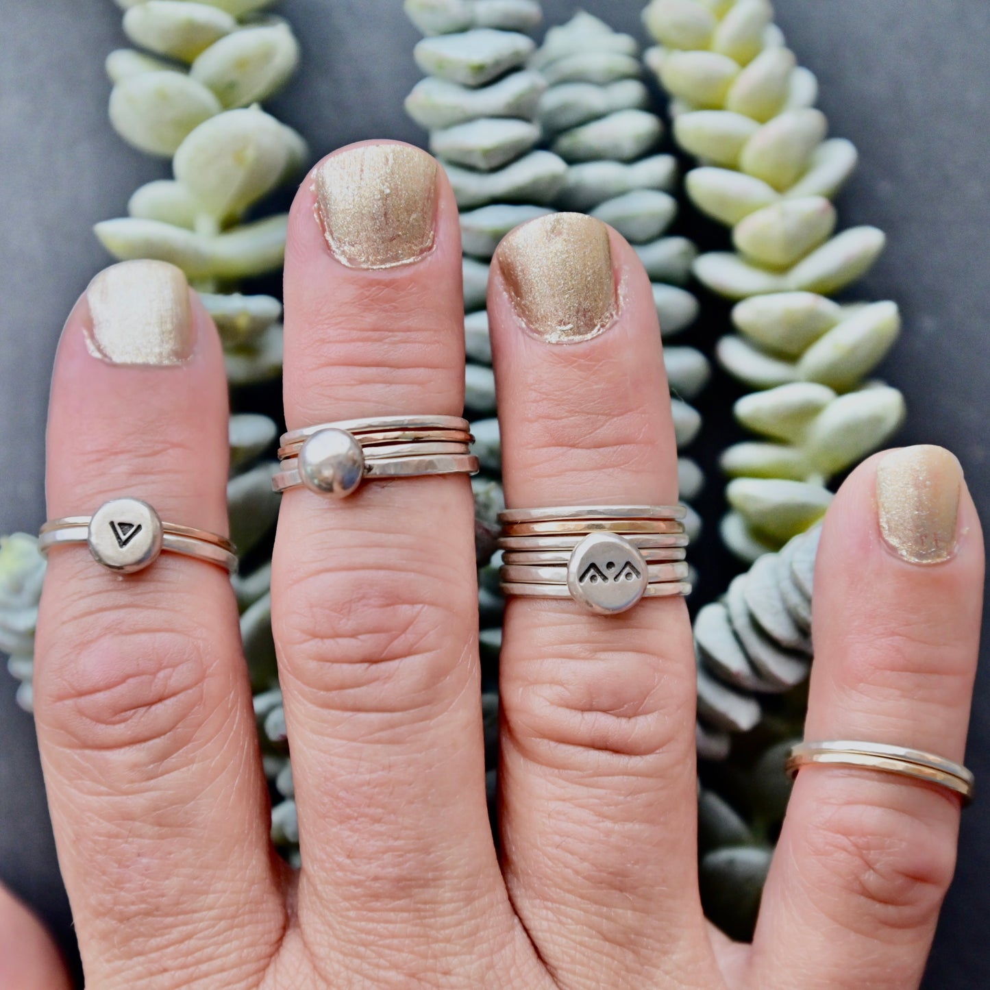 Hand wearing multiple silver and gold rings with a gray background and succulent plants.