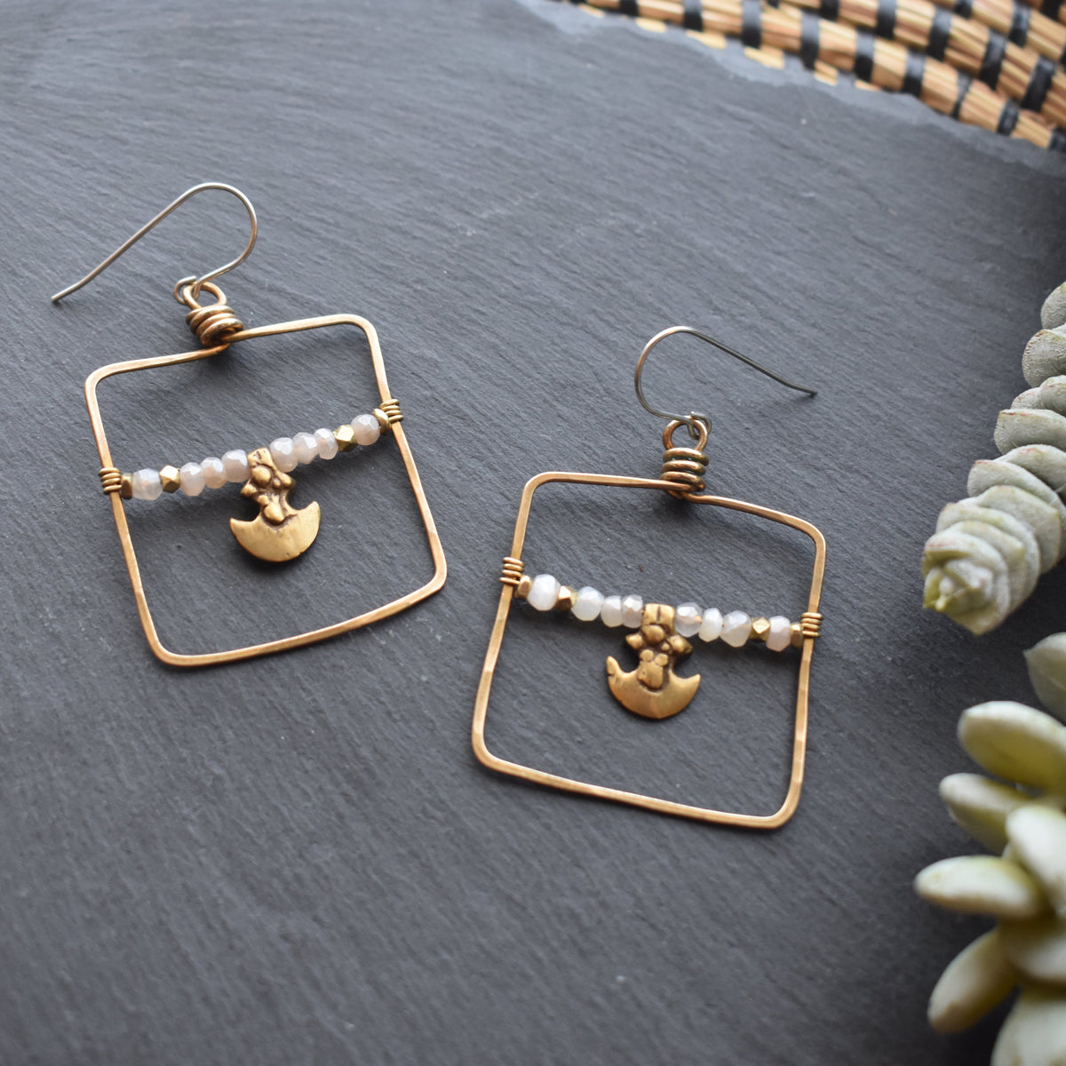 A pair of handmade gold wire dangle earrings with small white moonstone beads and a gold anchor charm, displayed on a dark slate surface next to a green succulent.